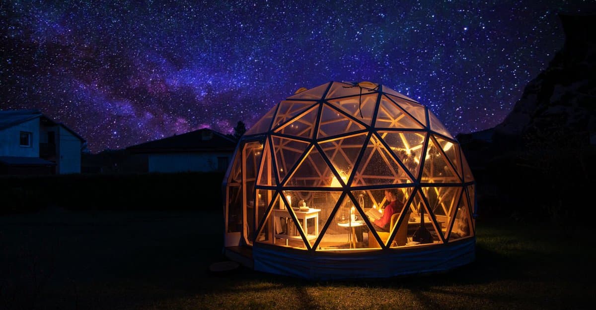 A geodesic dome tent is illuminated from within, revealing a person sitting at a table inside, set against a backdrop of a star-filled night sky and distant houses.