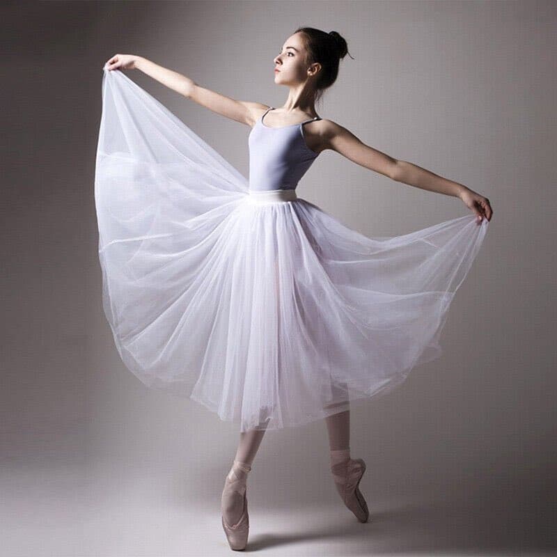 A young ballerina in a white leotard and tutu stands on pointe against a plain gray studio background, holding the edges of her flowing skirt with both arms extended.