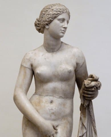 A photograph of a classical marble statue depicting a nude female figure with her hair in a bun, holding a draped cloth in her left hand.