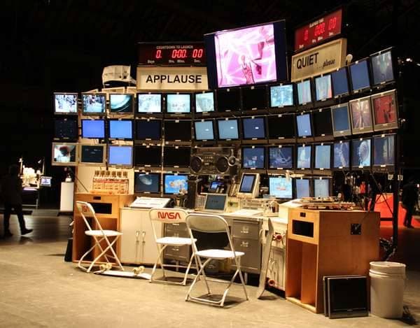 A photograph of an art installation resembling a NASA mission control room, featuring a wall of numerous television monitors, control panels, and chairs. The setup includes signs with the words "APPLAUSE" and "QUIET," along with digital displays showing numbers and the word "COUNTDOWN." The scene is set in a dark exhibition space with a few people visible in the background.