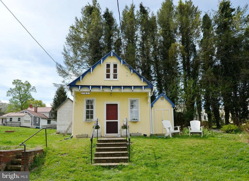 A small, yellow Victorian-style house with blue trim and a red front door sits on a grassy lawn, with a set of concrete steps leading up to the entrance. The house is surrounded by tall evergreen trees and other buildings in the background, with two white Adirondack chairs placed on the right side of the yard.