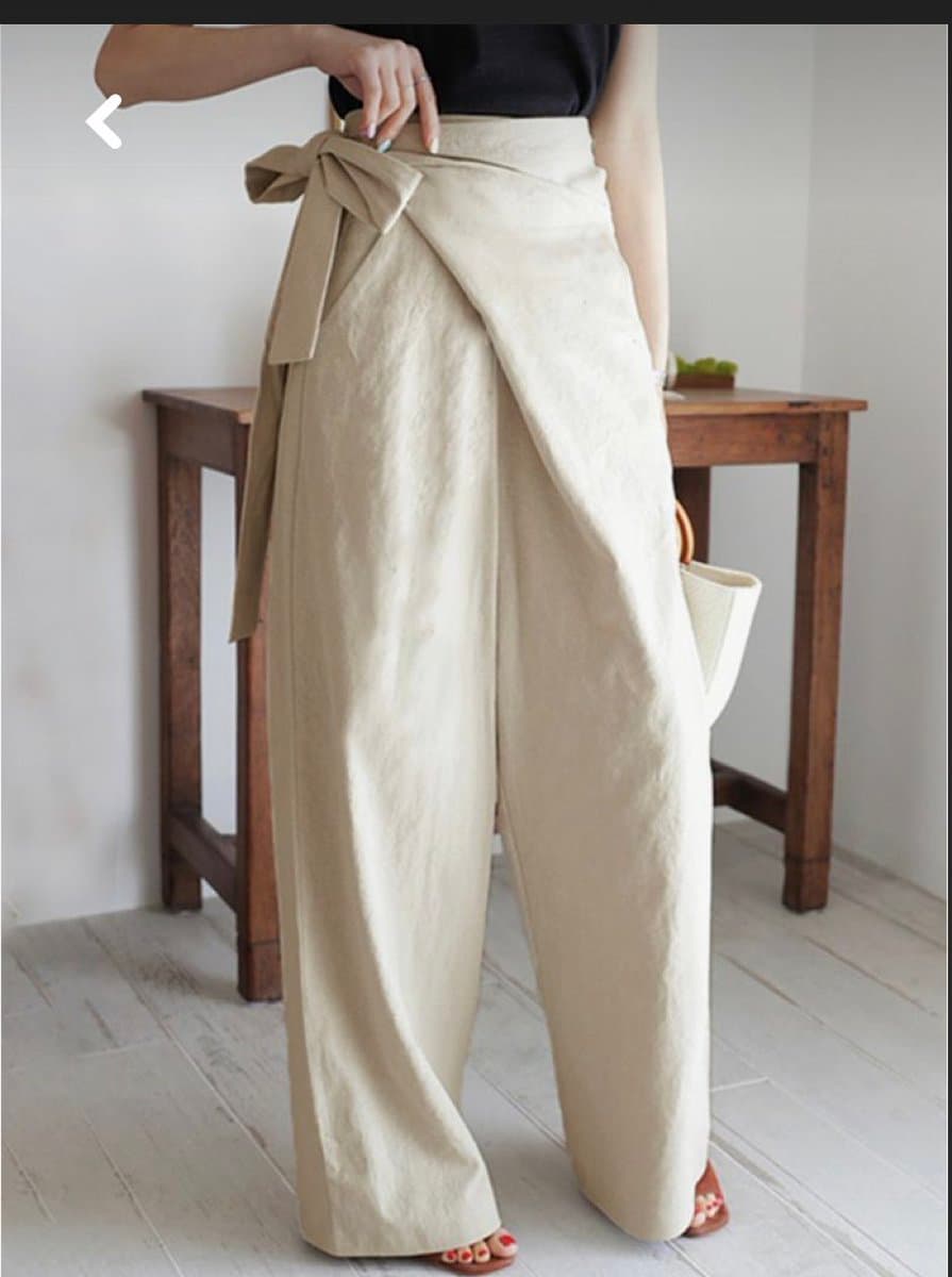 A screenshot of a fashion product photo showing a person from the neck down wearing a black top and wide-leg, cream-colored linen pants with a large bow at the waist. The person is standing on a light wood floor in front of a wooden table, holding a white woven bag.