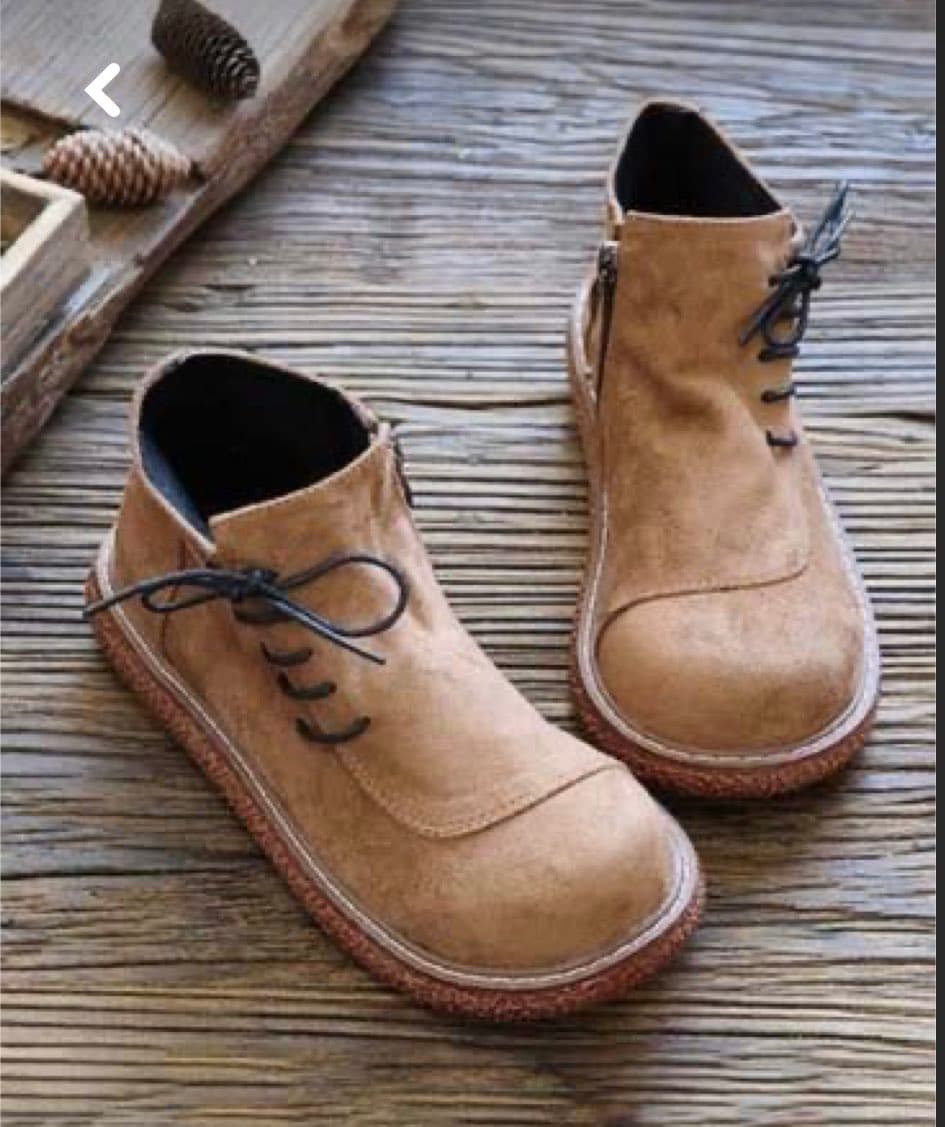 A pair of tan suede ankle boots with black laces and cork soles resting on a weathered wooden surface. A wooden tray with pinecones is visible in the upper left corner.