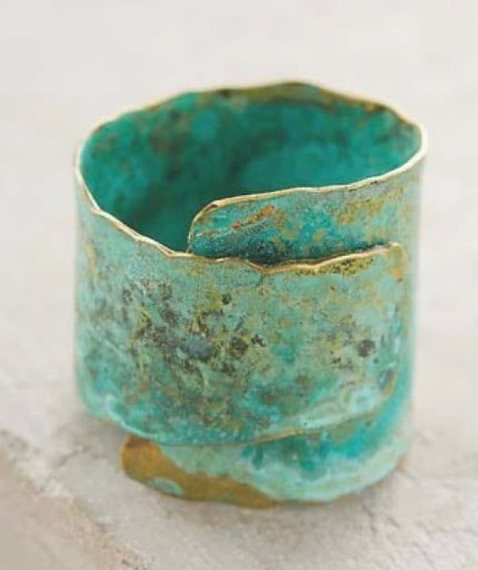 A close-up photograph of a wide, open cuff bracelet with a textured, oxidized turquoise and gold finish, resting on a light-colored surface. The piece has a rustic, handcrafted appearance with an irregular, organic shape.