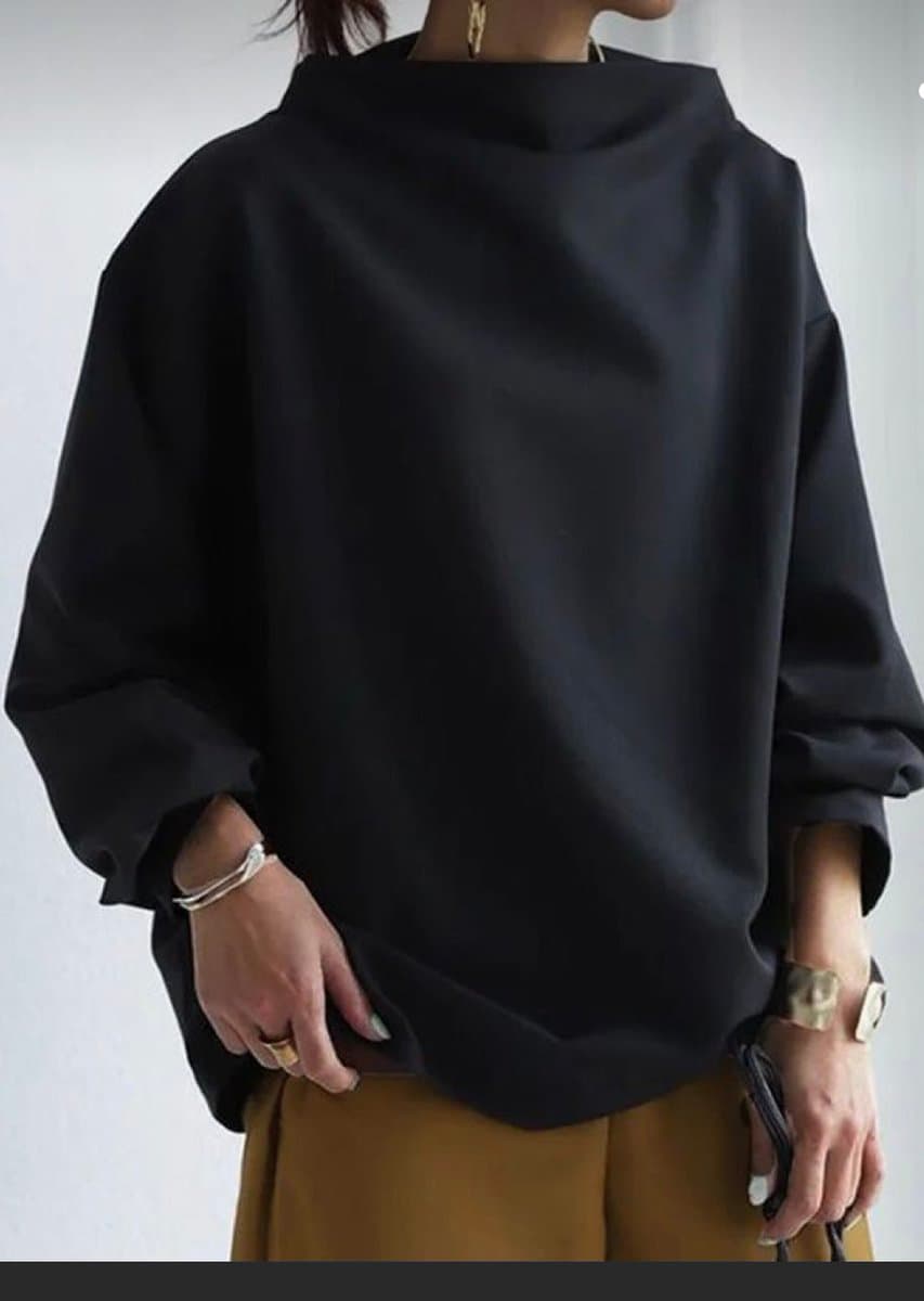 A person is shown from the neck down wearing a loose, oversized black top with a high collar and wide sleeves. They are wearing mustard-colored pants and gold-toned jewelry, including bracelets, rings, and a watch.