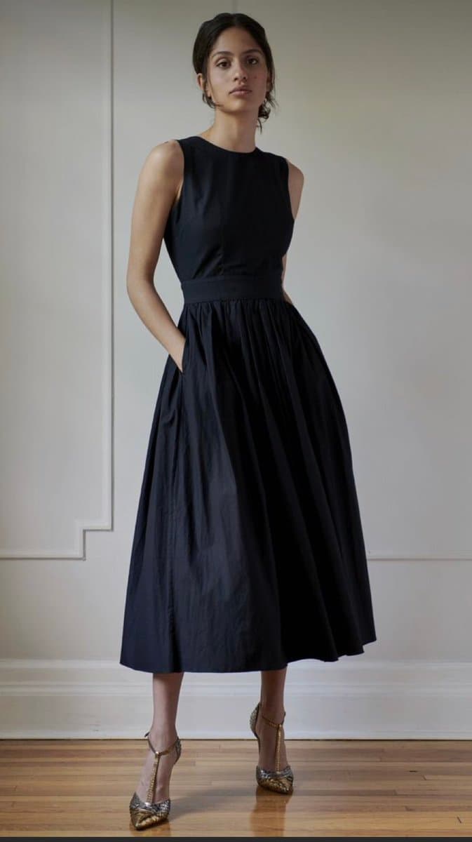 A full-length studio photograph of a woman standing against a white paneled wall, wearing a sleeveless, high-necked black midi dress and gold metallic heels.