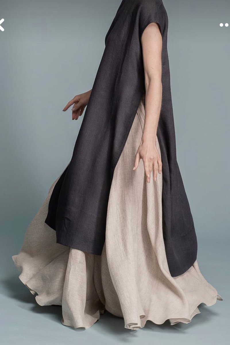 A person is shown from the shoulders down, wearing a long, dark gray sleeveless tunic over a flowing, light beige maxi skirt. The outfit is photographed against a plain, neutral gray background, highlighting the drape and texture of the garments.