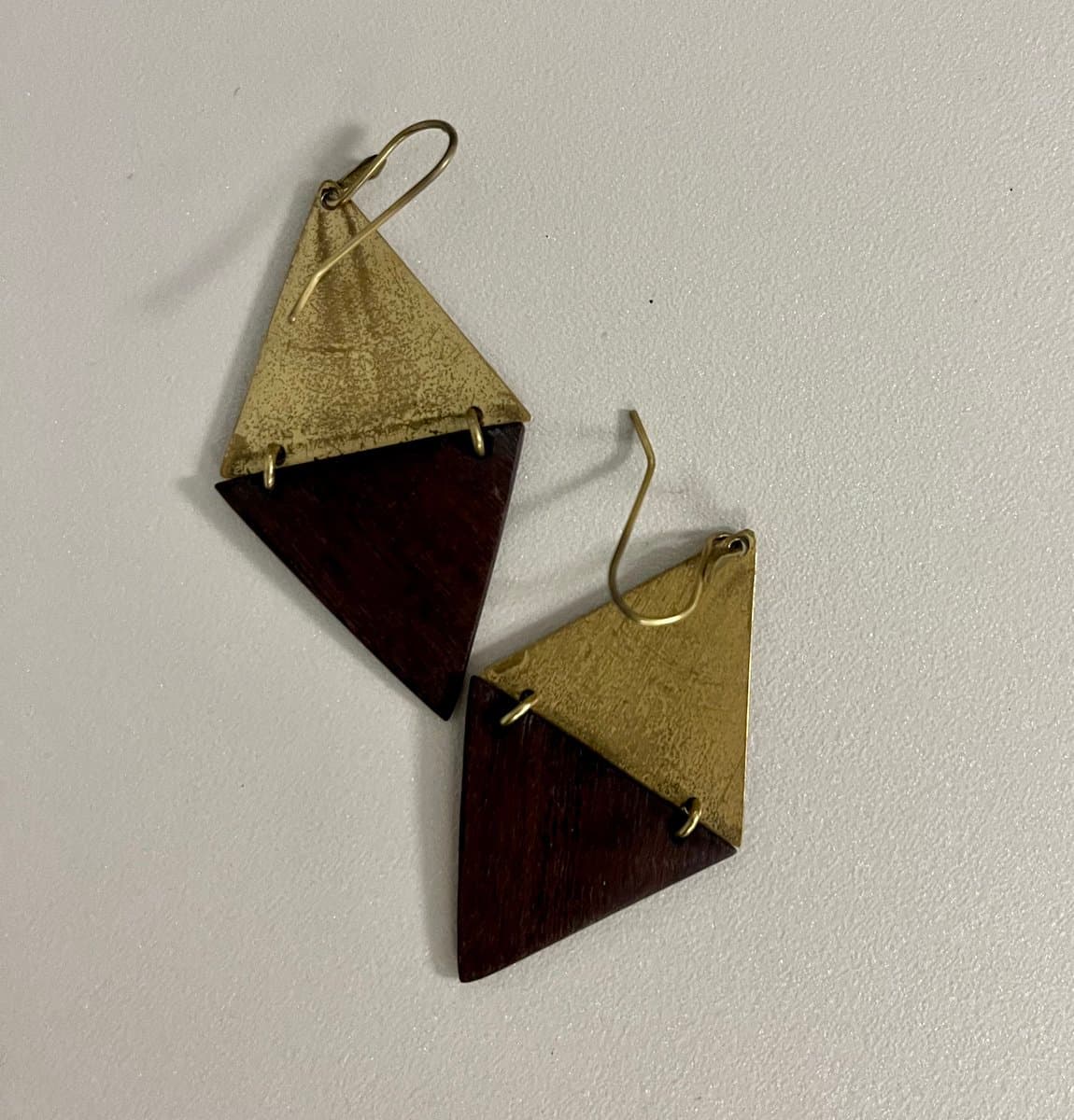 A close-up photograph of a pair of handmade, geometric earrings lying on a white surface. Each earring consists of a triangular gold-colored metal piece attached to a dark wood triangle, connected by small gold loops and a wire hook.