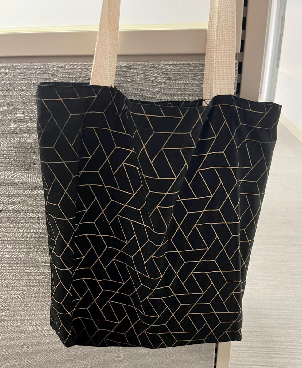 A black tote bag with a white geometric line pattern hangs from its white fabric handles. The bag is suspended in front of a textured gray wall and a window frame.