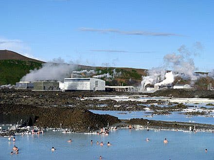 A scenic outdoor photograph of a geothermal lagoon with people swimming in the blue water, set against a backdrop of steam rising from a geothermal power plant and green hills. The image appears to be a standard landscape photo rather than a text-bearing graphic.