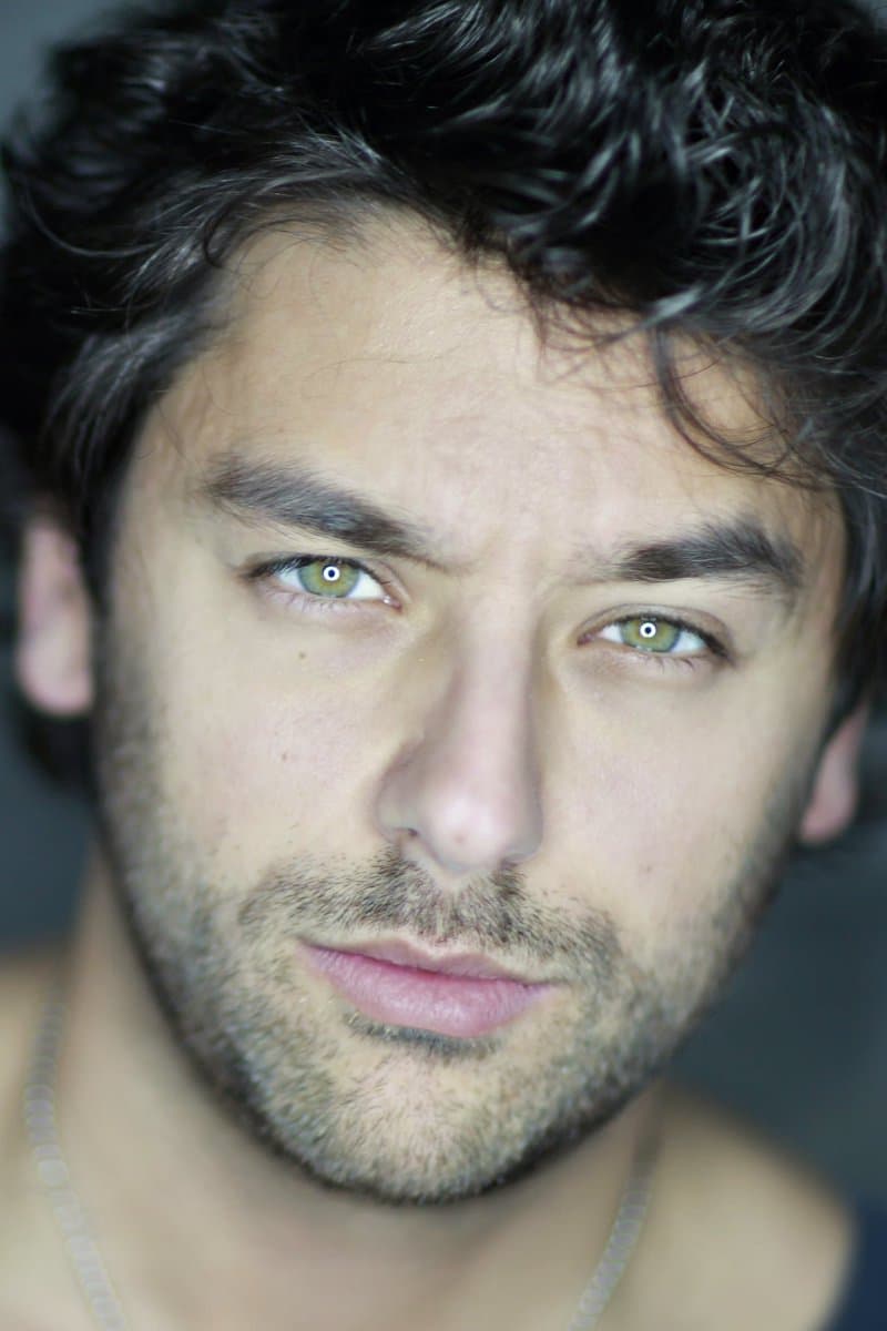 A close-up portrait of a man with dark, wavy hair and light-colored eyes, looking directly at the camera. He has light facial stubble and is wearing a thin chain necklace.