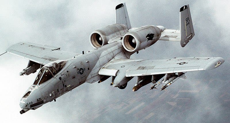 A photograph of a gray A-10 Thunderbolt II military aircraft flying in the sky, equipped with external weapons and featuring twin vertical stabilizers and rear-mounted engines. The image is a standard color photograph of the plane in flight.