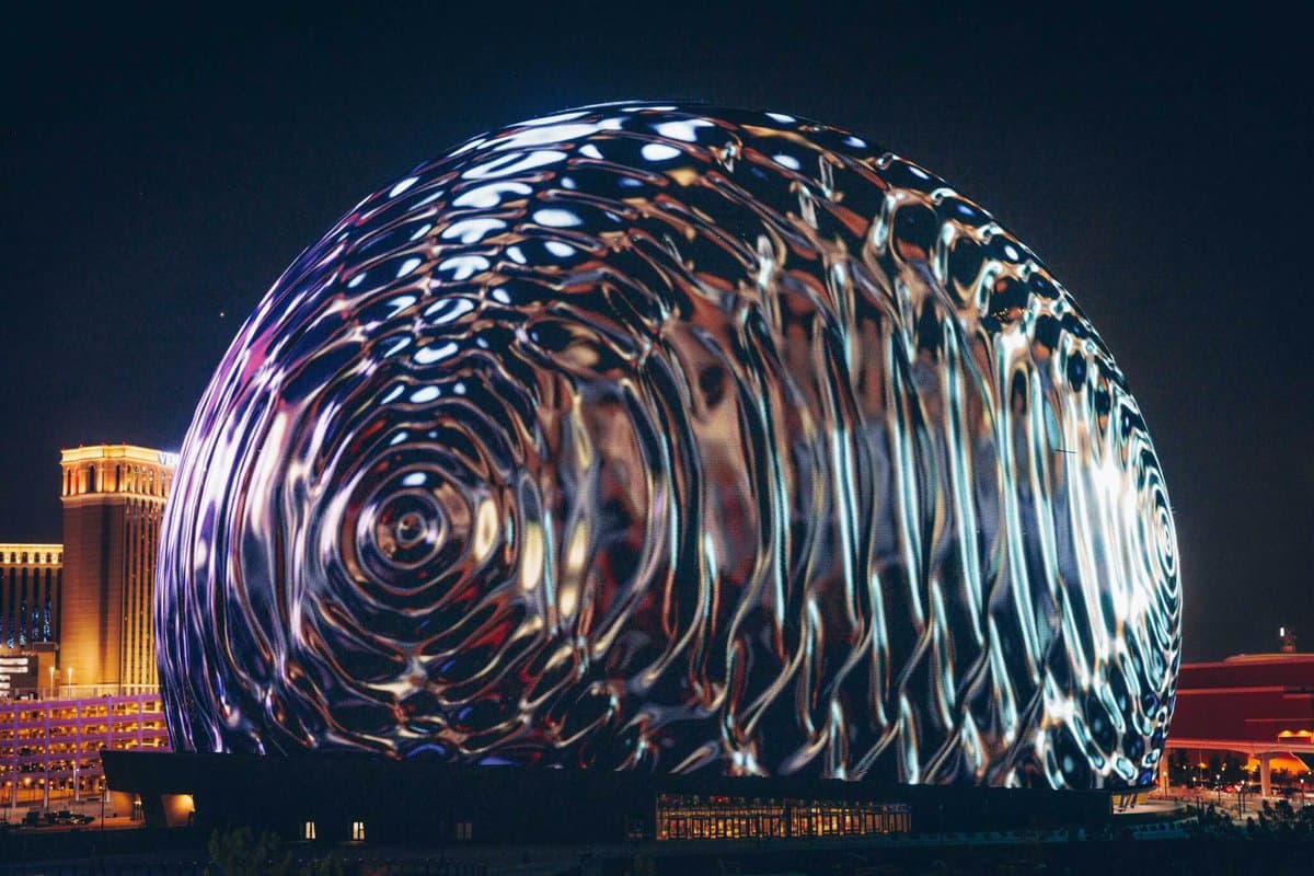 A nighttime photograph of a large, reflective metallic sphere with a rippled surface, illuminated by colorful lights against a dark sky. In the background, the illuminated facade of a large hotel or casino building is visible on the left.