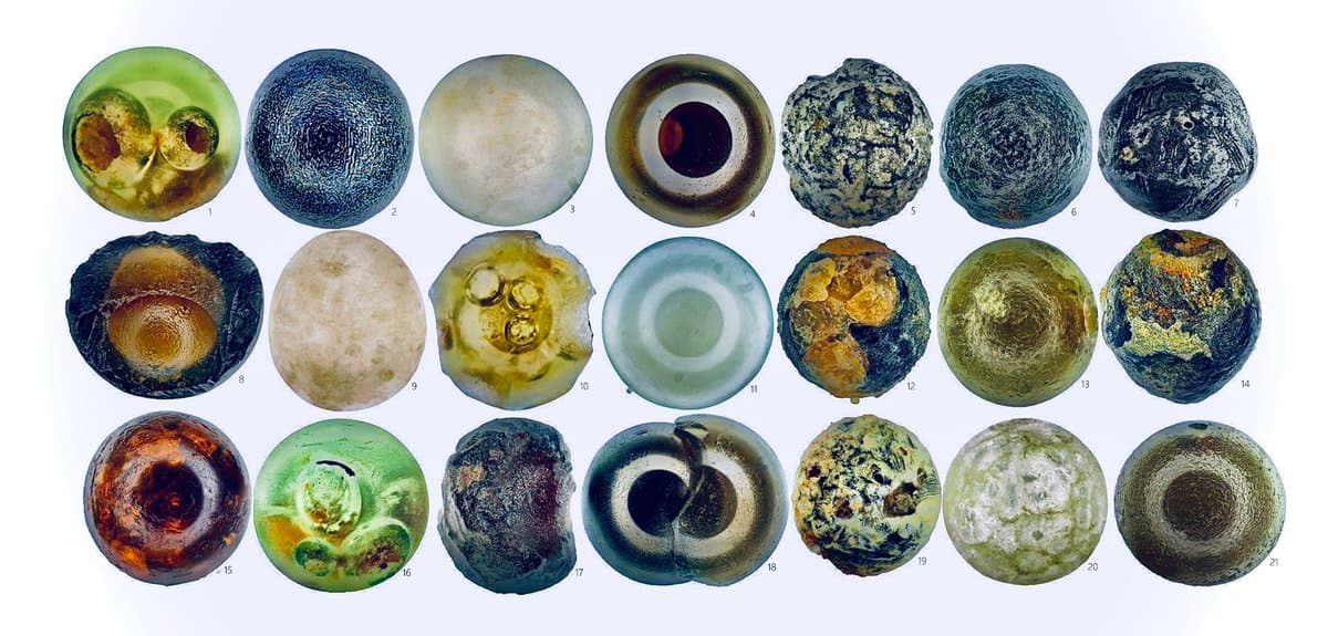 A grid of 21 numbered, circular microscopic images showing various colorful and textured Antarctic micrometeorites against a white background. The specimens display a wide range of colors, including green, blue, brown, and white, with diverse internal structures and surface patterns.