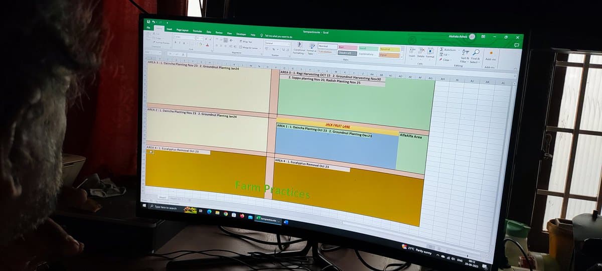 A photograph of a computer monitor displaying a Microsoft Excel spreadsheet used to plan farm layouts. The spreadsheet features colored cells representing different areas with text detailing planting and harvesting schedules, such as 'AREA 1: 1. Daincha Planting Nov 23' and 'JACK FRUIT LANE'.
