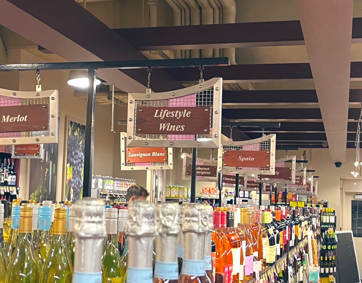 A photograph of a wine store aisle with rows of bottles and hanging wooden signs. The central sign reads "Lifestyle Wines," which is the subject of the post's humorous question. Other visible signs include "Merlot," "Savignon Blanc," and "Spain."