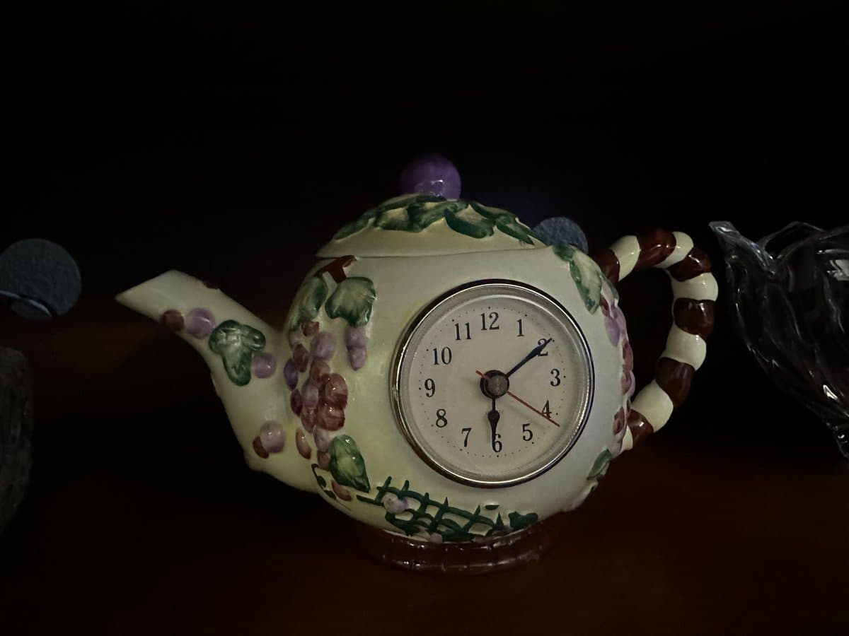 A ceramic teapot decorated with painted grapes and leaves, featuring a built-in analog clock on its side. The clock face displays the time as approximately 6:10.