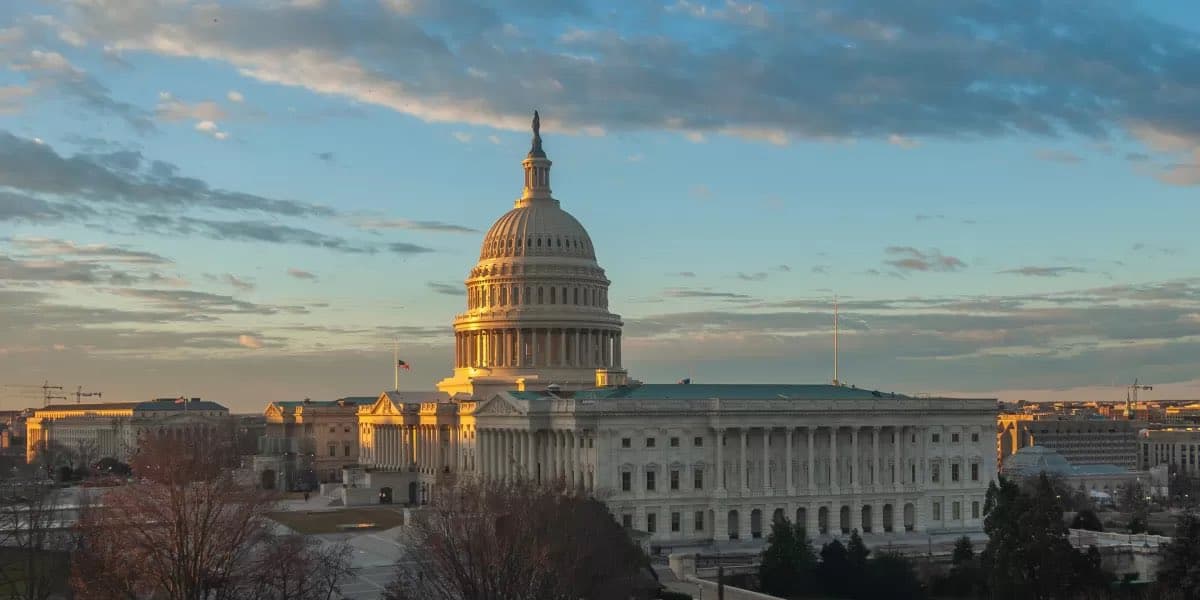 A wide-angle photograph of the United States Capitol building in Washington, D.C., taken during the golden hour of sunrise or sunset. The iconic dome and surrounding wings are illuminated by warm, low-angle sunlight against a blue sky with scattered clouds, with bare trees visible in the foreground.