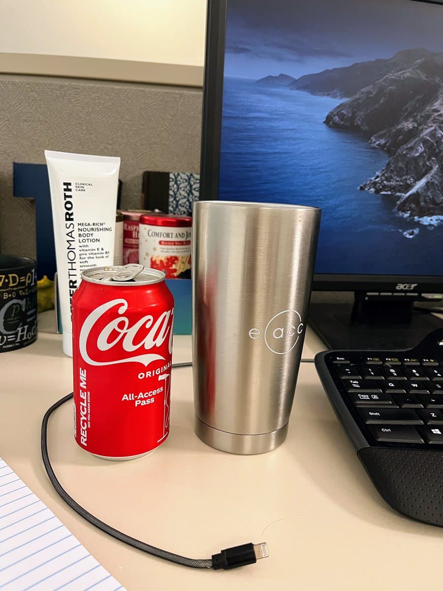 A desk setup featuring a red Coca-Cola can and a silver stainless steel tumbler with an 'e acc' logo, placed next to a computer monitor and keyboard. In the background, there is a tube of Peter Thomas Roth body lotion and a can of Comfort food.