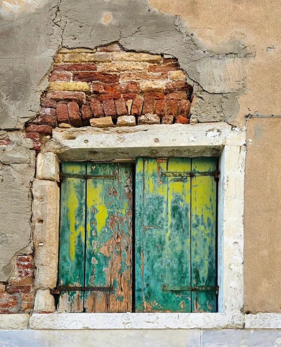 A close-up photograph of a weathered window with peeling green and yellow paint on the wooden shutters, set into a wall with exposed red brick and crumbling plaster.