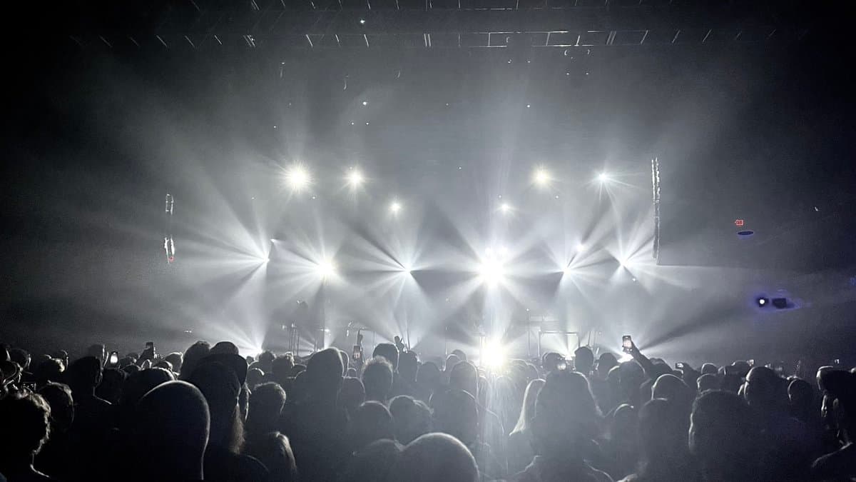 A black-and-white photograph of a concert crowd facing a stage illuminated by bright, radiating spotlights. The silhouettes of audience members are visible in the foreground, with some holding up phones to record the performance.