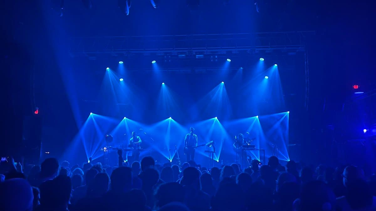 A wide shot of a live music performance on a stage bathed in intense blue light, with beams from overhead spotlights creating a dramatic atmosphere. A band is visible on stage in front of a large audience, whose silhouettes fill the foreground.