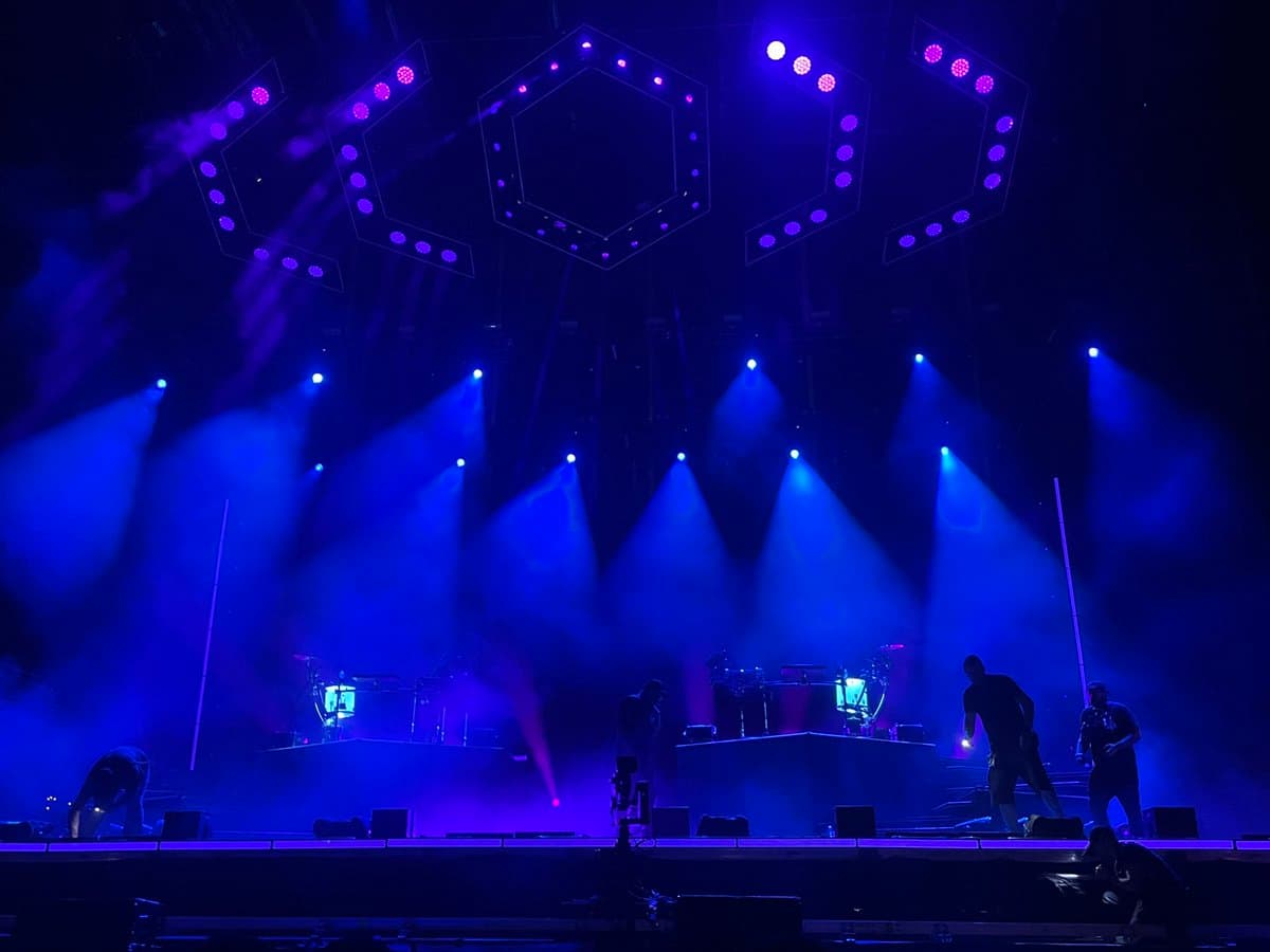 A wide shot of a concert stage bathed in blue and purple light, featuring silhouetted performers and a drum kit on a raised platform. Overhead, large hexagonal light rigs with purple bulbs hang above the stage, while bright spotlights cut through the atmospheric haze.