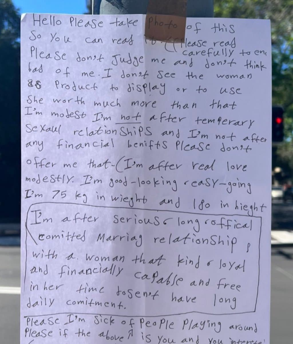 A photograph of a handwritten sign on a white sheet of paper, taped to a pole in an outdoor setting. The sign is a personal advertisement seeking a serious, long-term relationship and marriage, stating the author's physical attributes and preferences.