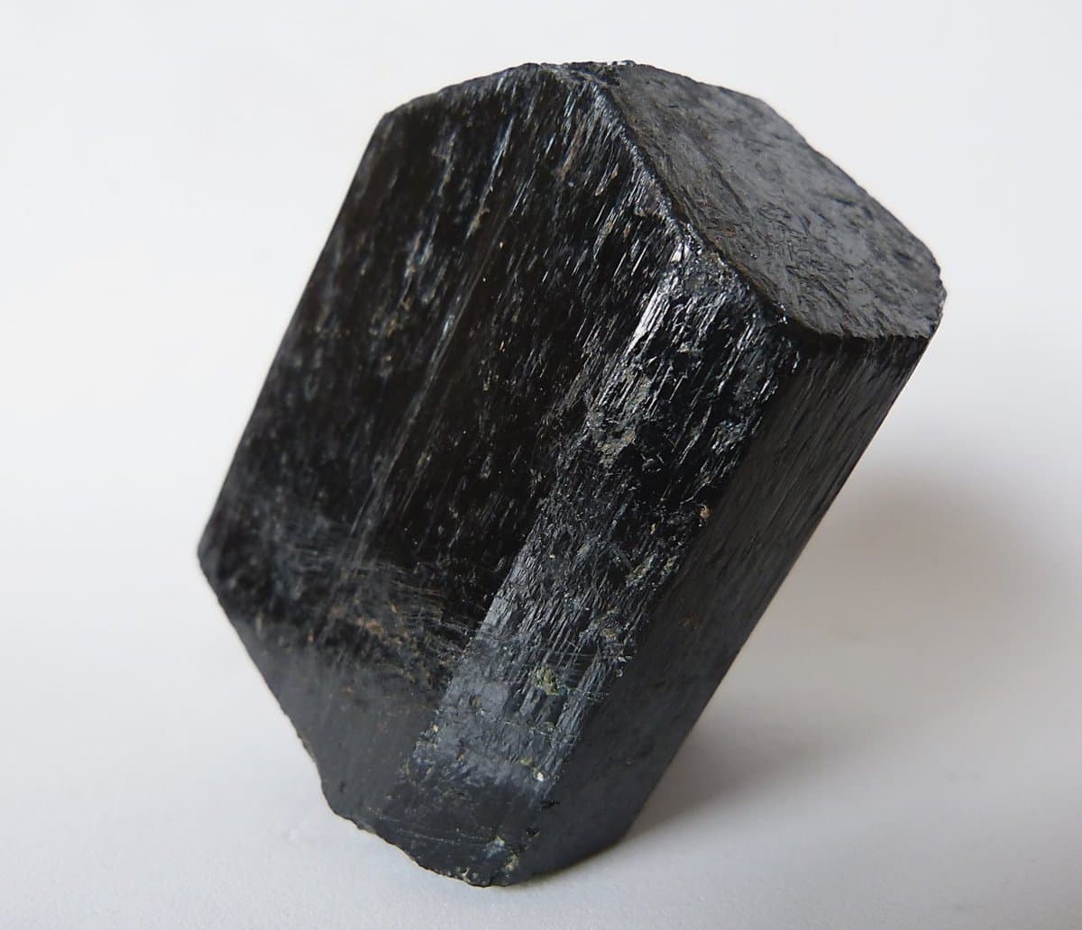 A close-up photograph of a large, dark, hexagonal crystal with a striated surface texture, set against a plain white background.