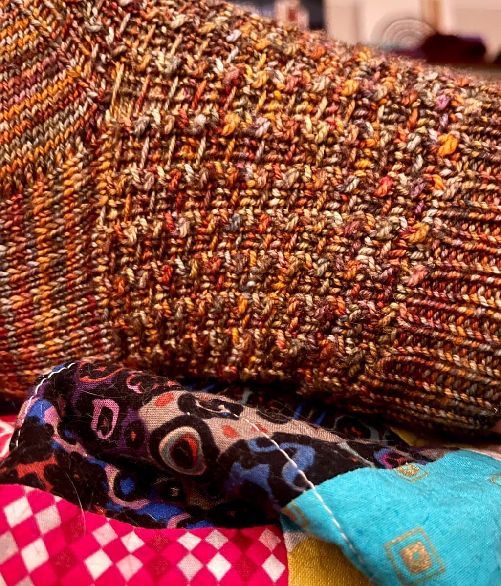 A close-up photograph of a folded, multicolored hand-knitted item, likely a pair of socks, resting on a pile of patterned fabrics. The knit features a textured stitch in warm autumnal tones of brown, orange, and red.