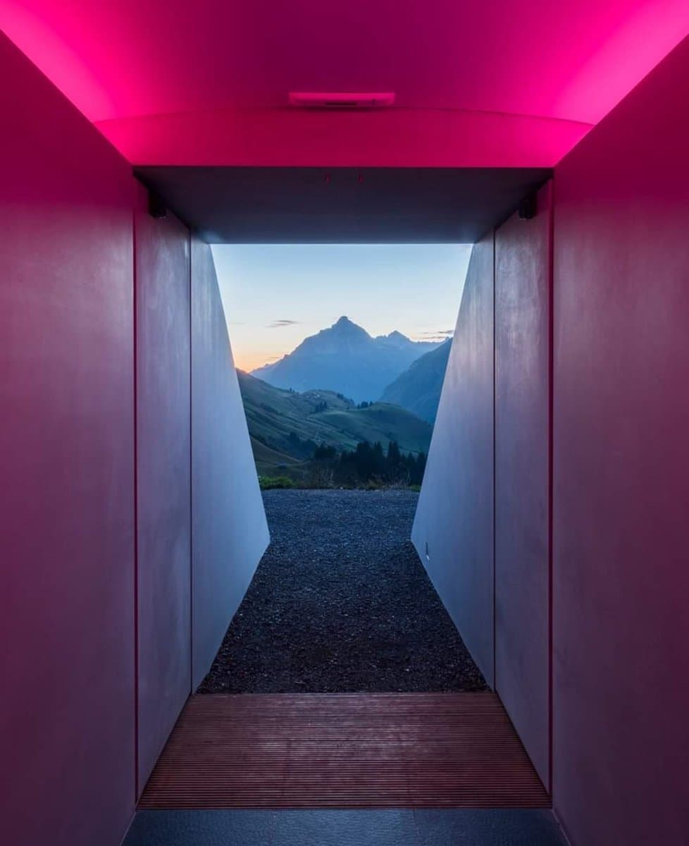 A photograph of a James Turrell light installation, showing a narrow interior space with a glowing pink ceiling that frames a view of a mountain landscape at dusk. The perspective leads from a wooden floor through a dark gravel path toward a distant, snow-capped peak.