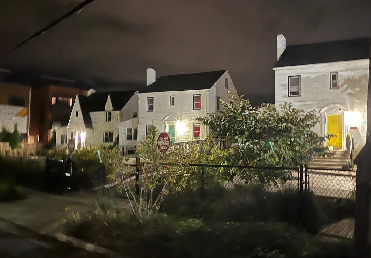 A nighttime photograph of a row of white residential houses with lit windows, viewed from across a street with a chain-link fence and bushes in the foreground.