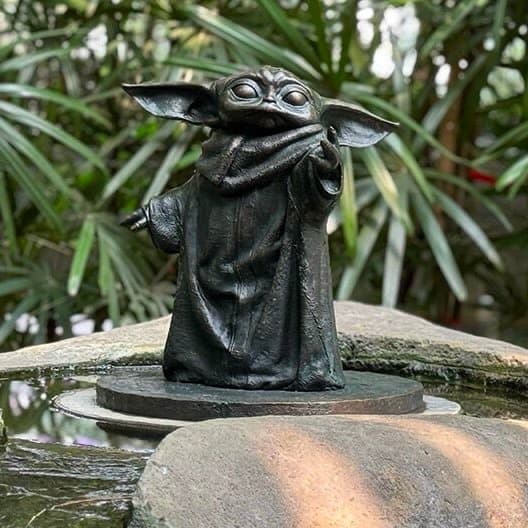 A small, dark-colored figurine of the character Baby Yoda (Grogu) from Star Wars, shown standing on a stone ledge with its hand near its mouth. The background features blurred green foliage, suggesting an outdoor garden setting.