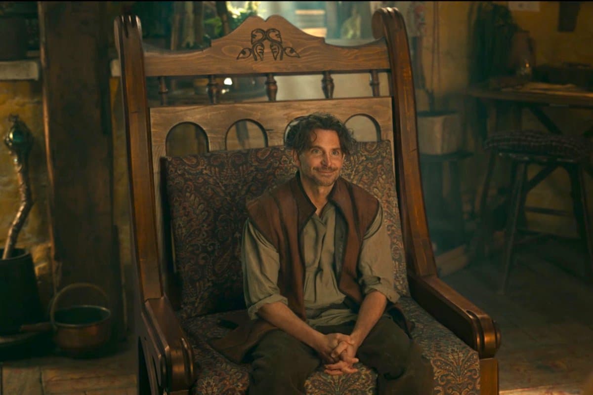 A still from a film showing a man with dark hair and a mustache sitting in a large, ornate wooden chair with patterned upholstery. He is wearing a light-colored shirt and a brown vest, with his hands clasped in his lap, set against a dimly lit, rustic interior background.