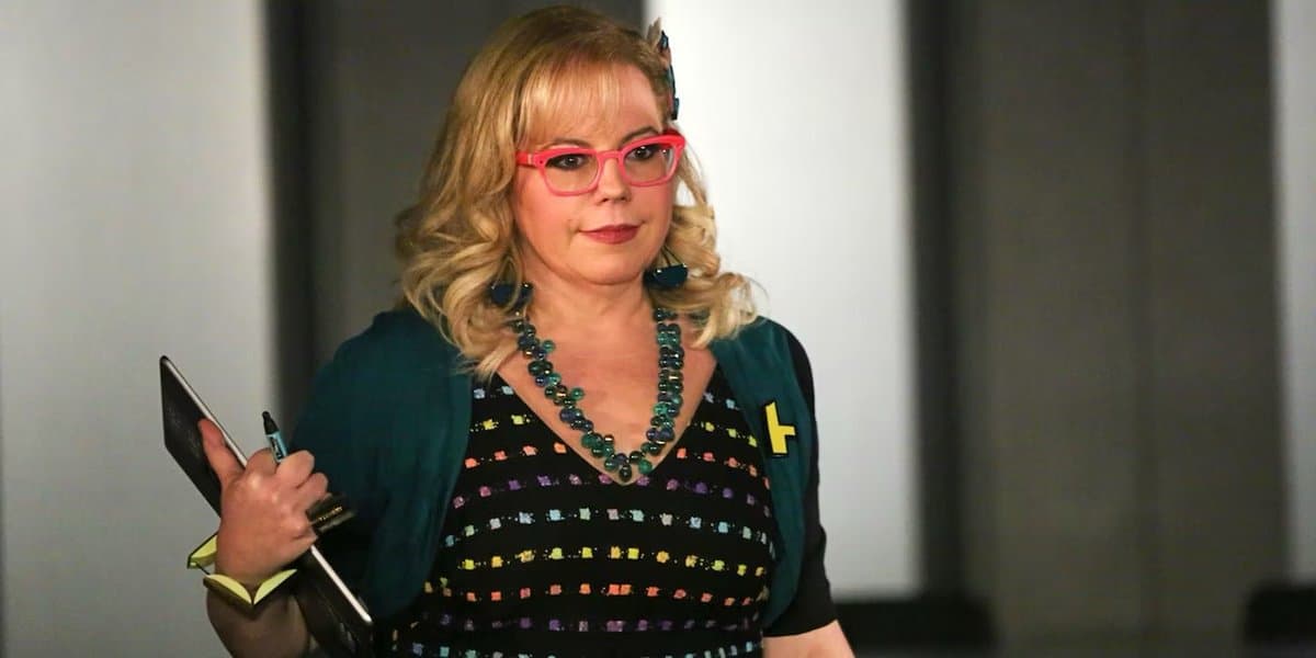 A woman with blonde hair, wearing bright pink glasses, a beaded necklace, and a patterned top, holds a notebook and pen while looking toward the camera.