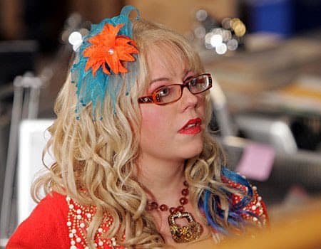 A woman with curly blonde hair, red glasses, and a colorful orange and blue flower headband is shown in profile, wearing a red beaded top and a necklace. She appears to be in an office or studio setting with blurred equipment in the background. The image is a photograph, likely a screenshot from a video or TV show.