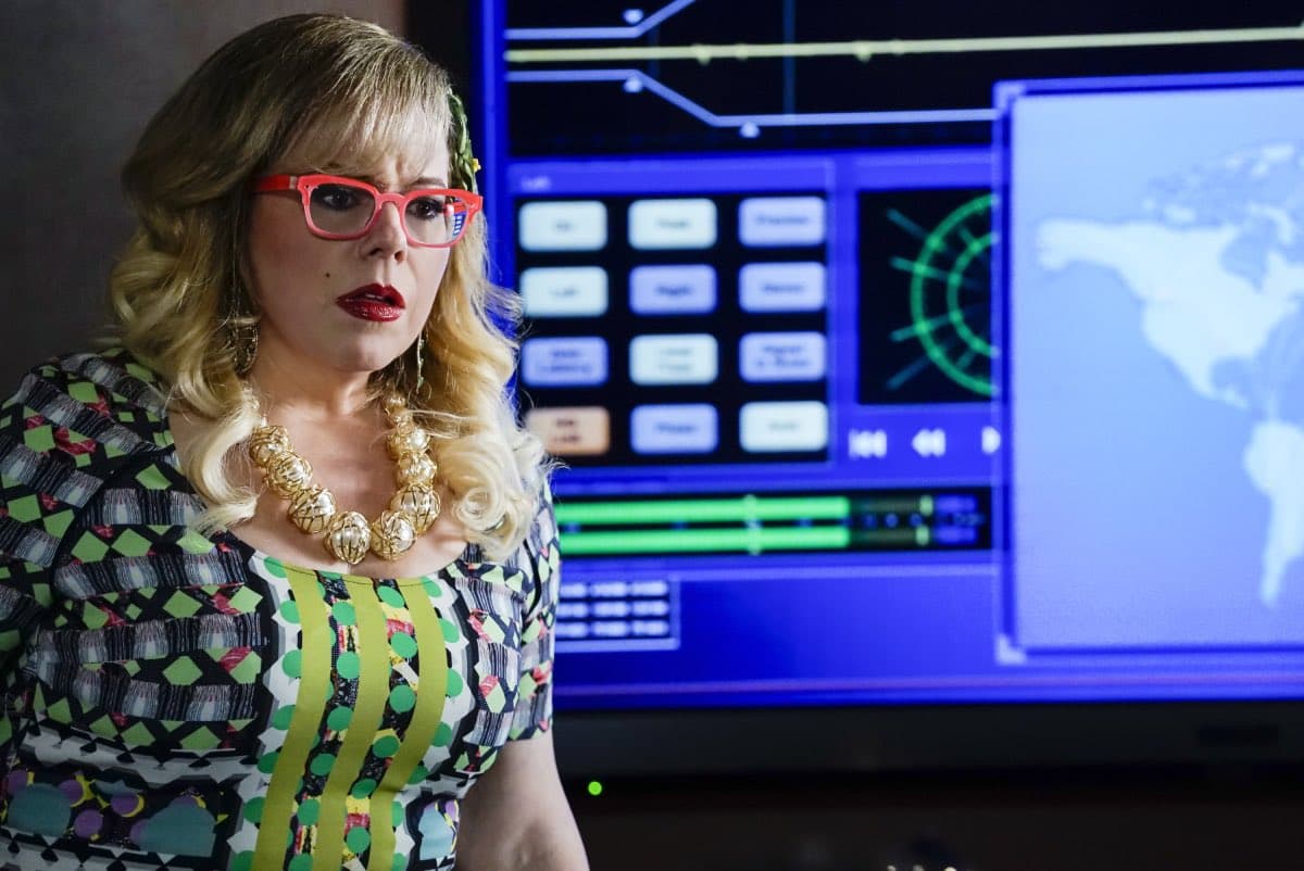 A woman with blonde hair, red glasses, and a colorful patterned dress stands in front of a large blue digital screen displaying a world map and various interface elements.