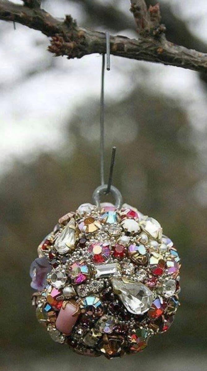 A close-up photograph of a spherical ornament covered in multicolored crystals and beads, hanging from a bare tree branch by a thin wire.