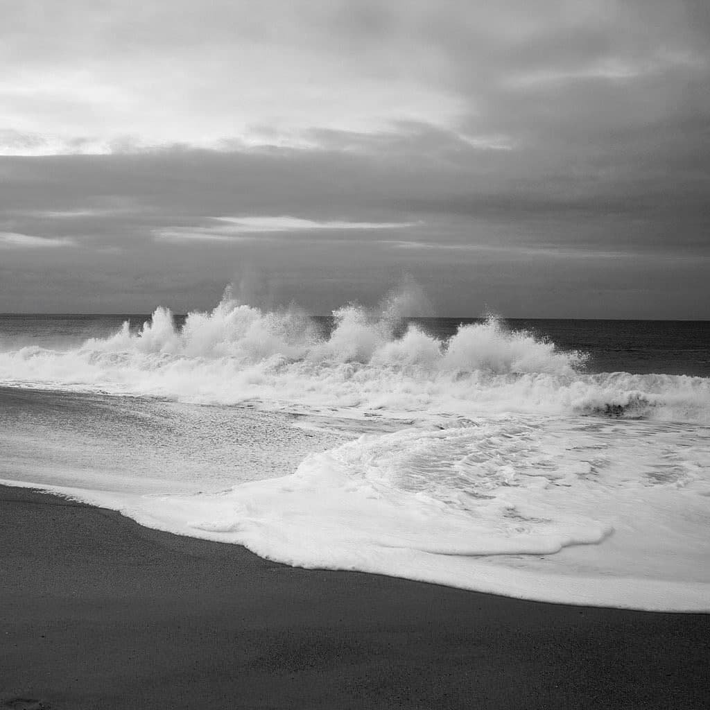 A black-and-white photograph of a beach with waves crashing onto the shore under a cloudy sky.