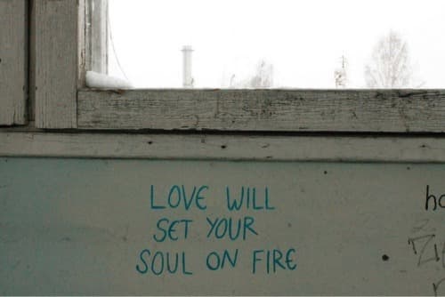 A photograph of a white wall beneath a window, with the phrase "LOVE WILL SET YOUR SOUL ON FIRE" written in blue marker. The window frame is weathered wood, and a snowy outdoor scene is visible through the glass.