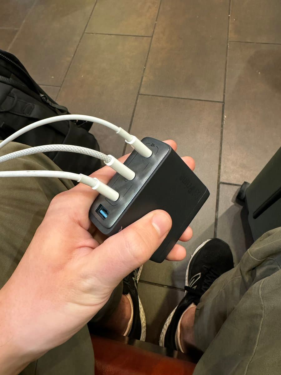 A first-person view of a person holding a black, multi-port USB charger with three white cables plugged in, while sitting on a tiled floor. The charger has a visible USB-A port and a USB-C port, and the person is wearing olive green pants and black sneakers.