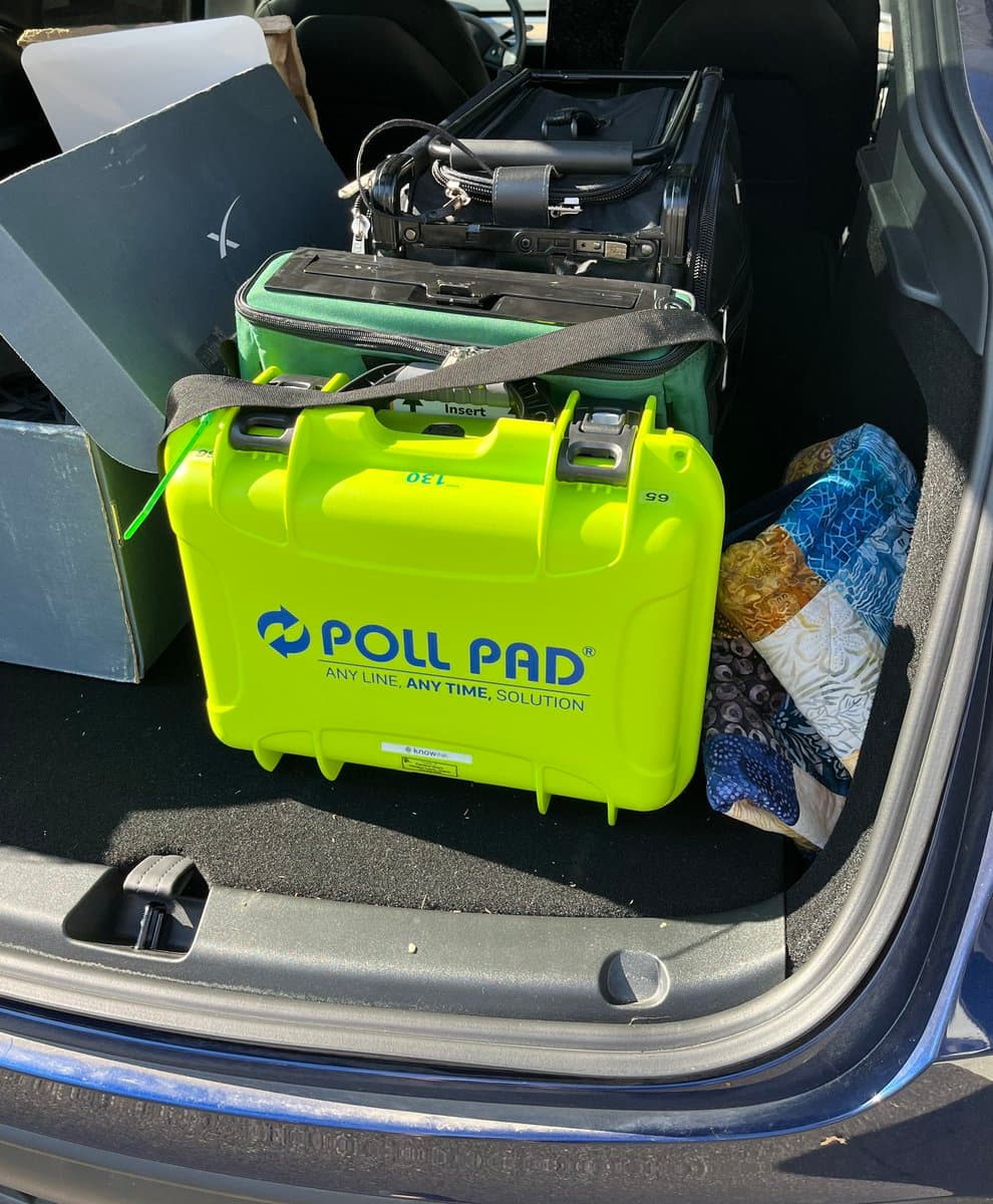 A photo of the open trunk of a car containing a bright green hard case labeled "POLL PAD" and other luggage. The case is positioned in the center of the trunk, surrounded by a black suitcase, a green bag, and a folded blanket.