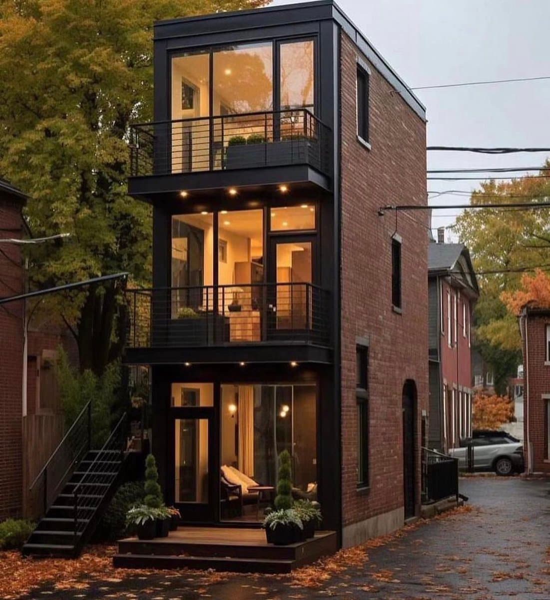 A photograph of a narrow, three-story brick townhouse with a modern black-framed glass and steel addition on the front facade. The house features large windows and balconies on each level, with warm interior lighting visible, and is situated on a residential street with autumn foliage.