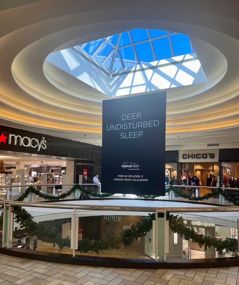 A photo taken inside a shopping mall looking up at a large, hanging advertisement banner for Tempur-Pedic. The banner is suspended from a circular skylight, with storefronts for Macy's and Chico's visible in the background.