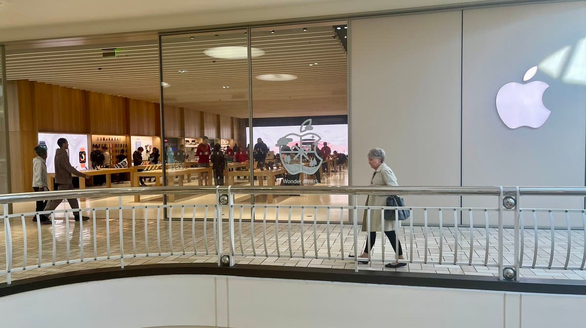 A photograph of the exterior of an Apple Store, showing a large white Apple logo on the wall and a glass storefront with wooden interior accents. A woman walks past the store on a tiled walkway with a metal railing.
