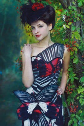 A woman with dark hair styled in an updo with a red flower accessory stands in a garden setting, wearing a black, white, and red patterned dress with a large bow at the waist. She is posing next to a tree with green leaves and red berries.