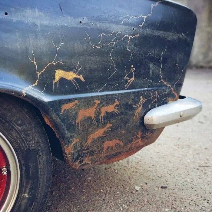 A close-up photograph of the rear fender of a dark blue vintage car, which features a custom airbrushed mural of deer and human figures in a style reminiscent of ancient cave paintings. The paint is heavily cracked and peeling, revealing rust underneath, and the car is equipped with a chrome bumper and a red wheel rim.