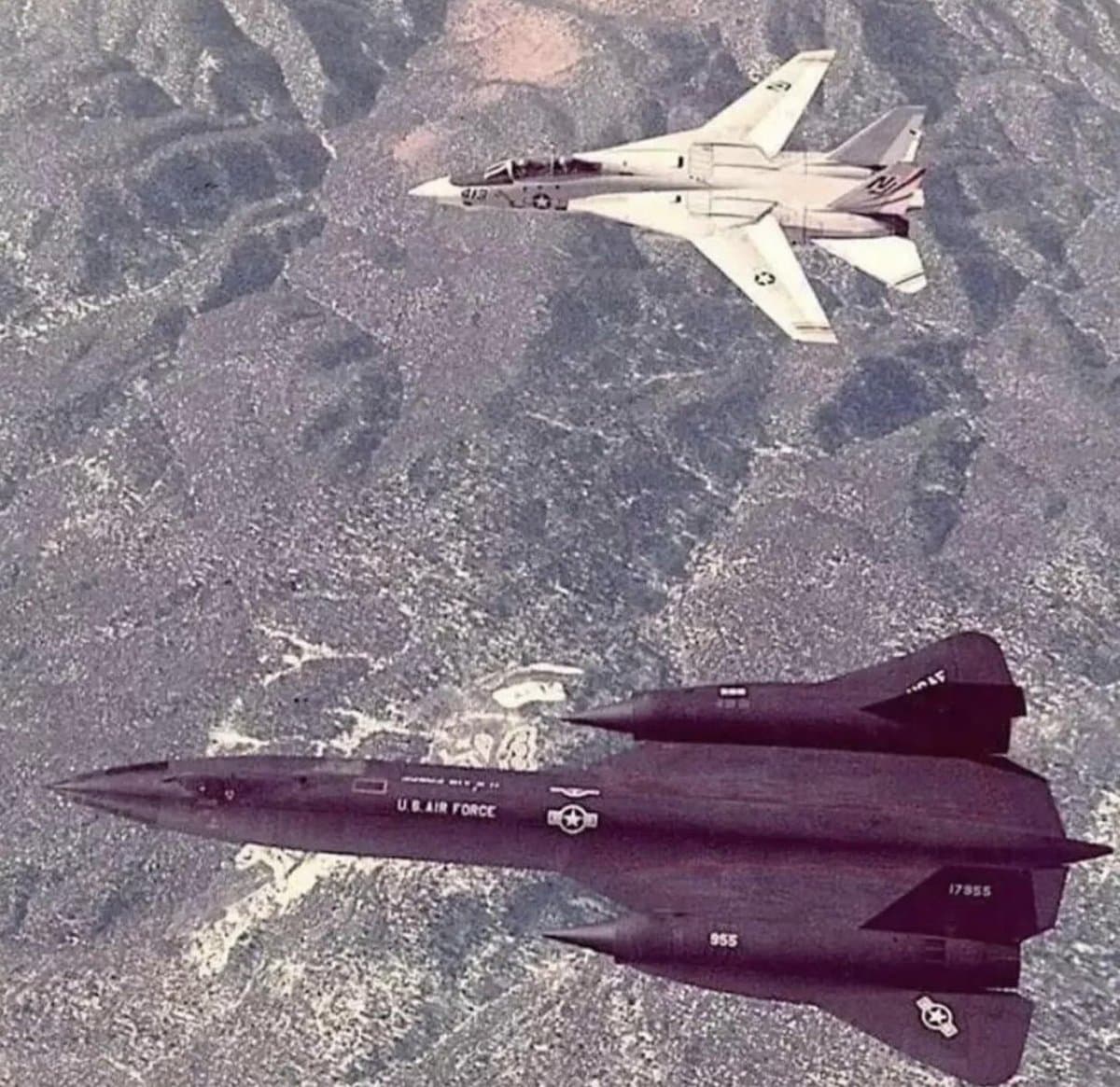 A photograph showing a black U.S. Air Force SR-71 Blackbird flying in formation with a white F-15 Eagle over a rugged, mountainous landscape.