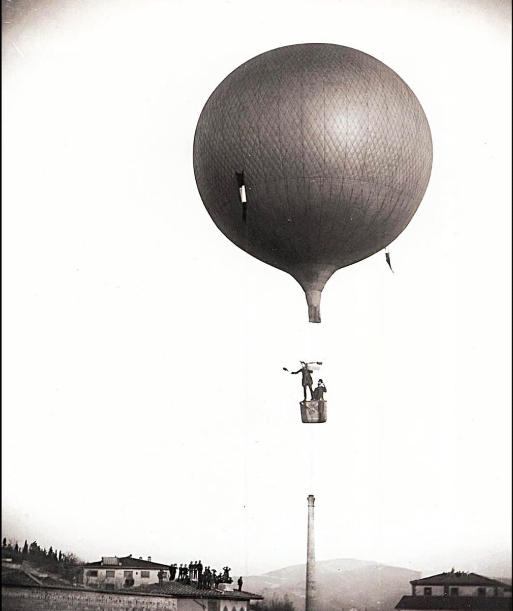 A vintage black-and-white photograph showing a large, spherical hot air balloon floating high in the sky above a town. A small basket suspended beneath the balloon contains two figures, one of whom is waving, while a crowd of onlookers gathers on the rooftops of buildings below.