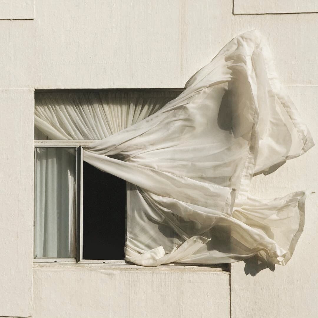A white curtain billows dramatically out of an open window on a light-colored building facade, caught in a strong gust of wind. The fabric is stretched and rippled, creating a dynamic, sculptural shape against the plain wall.